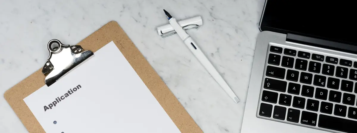An application form on a clipboard beside a pen and a laptop
