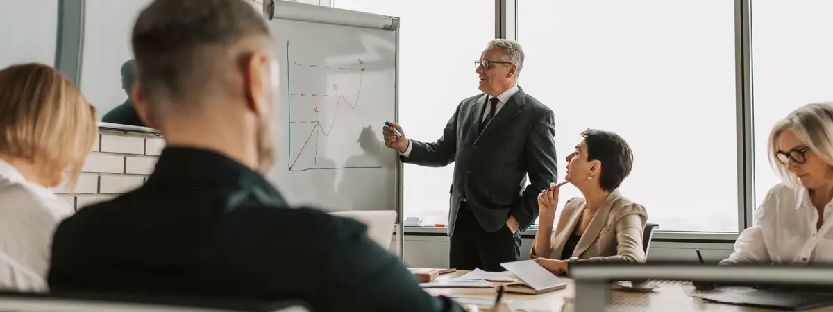 A consultant presenting charts at a whiteboard to colleagues around a table