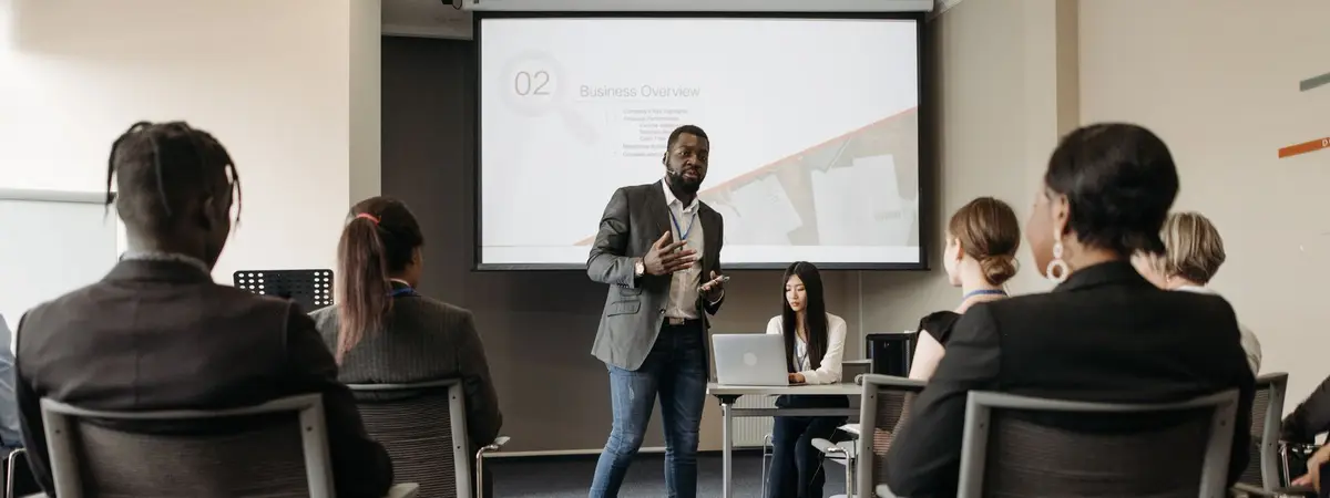 A speaker presenting a business overview slide to a seated group of professionals