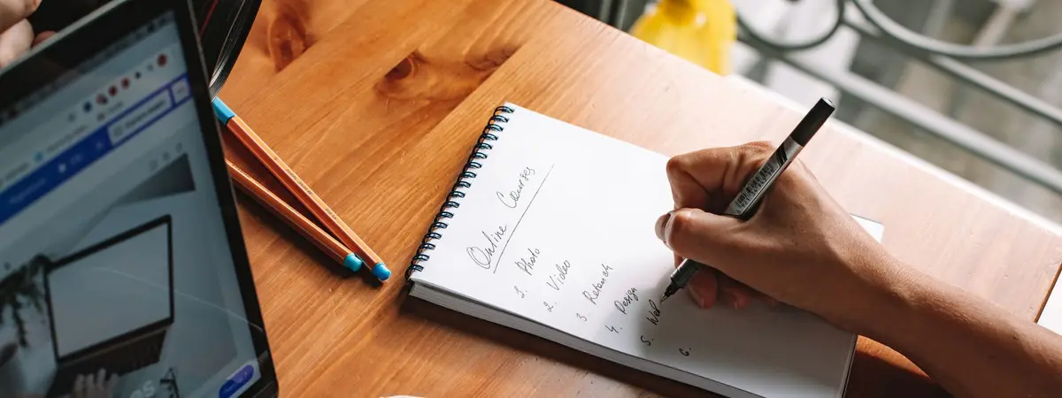 A person taking notes beside a laptop while researching online courses